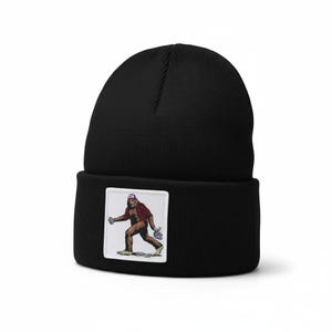 May include: Black beanie with a rectangular patch featuring a cartoon Bigfoot wearing a red and black plaid shirt and a baseball cap. The Bigfoot is holding a bouquet of purple flowers. The beanie is made of a soft, knitted material.