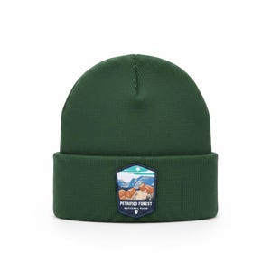 May include: A dark green knit beanie with a folded cuff. Centered on the cuff is a shield-shaped patch featuring a landscape illustration of Petrified Forest National Park with orange rocks, blue mountains, and the text Petrified Forest National Park.