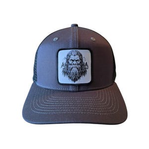 May include: A gray trucker hat with a black mesh back. The hat features a white patch with a black and white illustration of a Bigfoot's face.