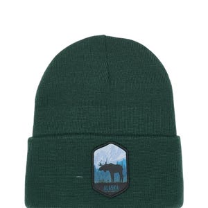 May include: A dark green knit beanie with a patch featuring a silhouette of a moose in front of a mountain range. The patch has the text "ALASKA" and "THE LAST FRONTIER" in white.