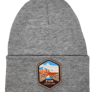 May include: Gray knitted beanie with a brown leather patch featuring a scenic view of Zion National Park and the text "Zion National Park".