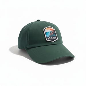May include: A dark green baseball cap featuring a shield-shaped patch on the front. The patch displays a landscape illustration of cliffs and water under a sun, with the text ACADIA NATIONAL PARK printed in white letters at the bottom.