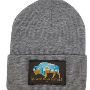 May include: Gray knitted beanie with a brown patch featuring a silhouette of a buffalo, mountains, and a sunrise. The patch also includes the text "National Park Service 1916".