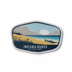 May include: An embroidered patch featuring a landscape of Indiana Dunes National Park with a sandy beach, blue water, and clouds in the sky. The text INDIANA DUNES NATIONAL PARK is stitched in white at the bottom of the oval-shaped patch.