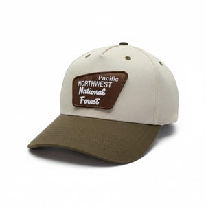 Op de afbeelding: Een crèmekleurige baseballpet met een olijfgroene klep en een bruine patch aan de voorzijde. De patch bevat de wit geborduurde tekst Pacific NORTHWEST National Forest. De pet heeft een knoop aan de bovenkant en bijpassende olijfgroene oogjes.