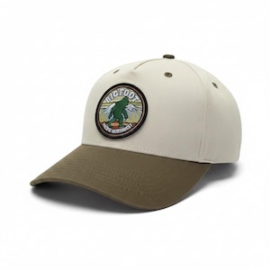 May include: A cream-coloured baseball cap with an olive green brim and a circular embroidered patch on the front. The patch features a green Bigfoot silhouette, mountain peaks, and the text "BIGFOOT PACIFIC NORTHWEST" in a circular border.