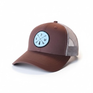 Può includere: Cappellino da camionista marrone con una toppa circolare azzurra con un design a bussola con le lettere "N", "P" e "W", e un albero e frecce. Il cappellino ha una parte anteriore e una visiera marroni e un retro in rete grigia.