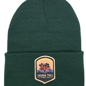 Op de afbeelding: Een donkergroene gebreide beanie met een patch met het logo van Joshua Tree National Park. Het logo is een kleurrijke illustratie van een Joshua Tree met een zonsondergang op de achtergrond.