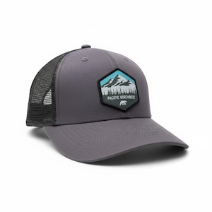 May include: Grey trucker cap with a black mesh back and a curved peak. The front displays a patch with a mountain scene, a bear, and the text "PACIFIC NORTHWEST". A fashionable accessory for outdoor activities.