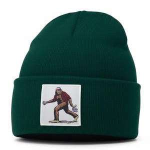 May include: A dark green knit beanie featuring a square white patch on the front cuff. The patch displays a brown, hairy creature wearing a red plaid shirt and a blue and white baseball cap, walking while holding purple flowers in one hand.
