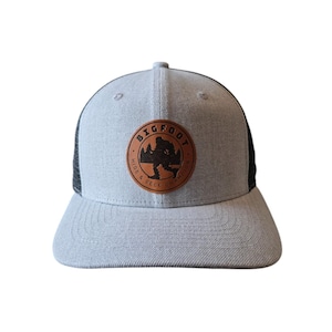 May include: A gray and black trucker hat with a brown leather patch that says "BIGFOOT HIDE & SEEK NATION".