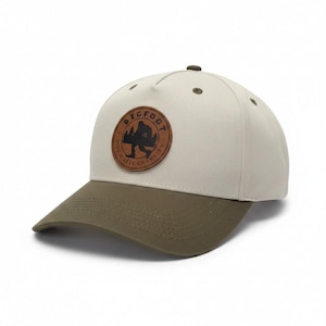 May include: A baseball cap with an off-white crown and an olive green brim. A circular brown leather patch is centered on the front, featuring a black silhouette of Bigfoot and the text "BIGFOOT HIDE & SEEK CHAMPION" embossed around the edge.