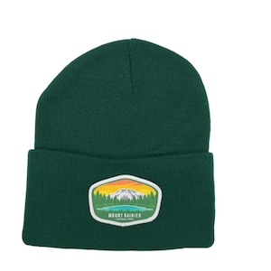 Puede incluir: Un gorro de punto verde oscuro con un parche blanco que presenta una ilustración colorida del Parque Nacional Mount Rainier.