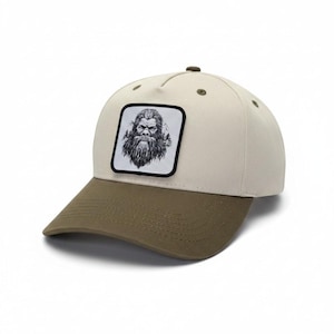 May include: A baseball cap featuring a cream-coloured crown and an olive green brim. A square patch is centred on the front, displaying a detailed black and white illustration of a bearded creature resembling a sasquatch amidst pine trees.