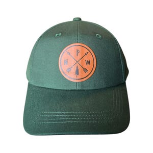 May include: A dark green baseball cap with a brown leather patch featuring a compass design with arrows pointing north, west, and south. The patch also includes a small pine tree graphic and the letters "P" and "W" inside the compass.