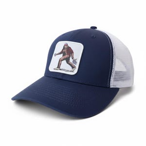 Op de afbeelding: Marineblauwe truckerpet met een witte mesh achterkant en een gebogen klep. De voorkant heeft een vierkante patch met een illustratie van Bigfoot. Bigfoot draagt een rood geruit overhemd en een rode hoed, met bloemen.