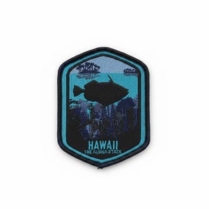 Puede incluir: Un parche bordado hexagonal que presenta la silueta de un pez negro nadando sobre una escena de arrecife de coral azul oscuro contra un fondo de agua azul claro. El texto HAWAII THE ALOHA STATE está bordado en azul claro en la parte inferior.