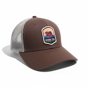 May include: A brown baseball cap with a mesh back and a curved brim. The front features a patch with a Joshua Tree National Park design, showcasing a desert landscape with a sunset. The text "JOSHUA TREE NATIONAL PARK" is visible.