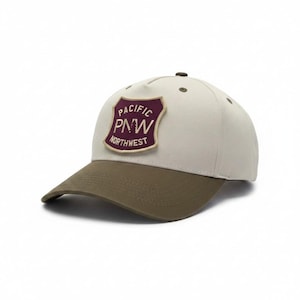 May include: A baseball cap with a light cream crown and an olive green brim. The front features a shield-shaped patch in dark maroon with a cream border. The text on the patch reads PACIFIC PNW NORTHWEST in cream-coloured capital letters.