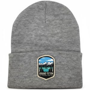 May include: A heather gray knit beanie featuring a folded cuff and a centered patch. The patch displays a graphic of the Grand Teton National Park with blue skies, white snow-capped mountains, green pine trees, and a lake, with the text GRAND TETON NATIONAL PARK.