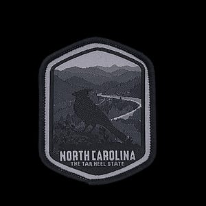 以下が含まれることがあります： 山と鳥のシルエットが刺繍された白黒のパッチ。画像の下に「NORTH CAROLINA THE TAR HEEL STATE」というテキストが印刷されています。