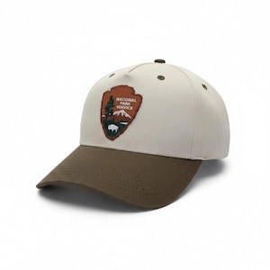 May include: A beige baseball cap with a dark brown brim and a brown embroidered patch on the front. The patch is shaped like an arrowhead and features the text NATIONAL PARK SERVICE, a green tree, a white mountain range, and a white bison silhouette.
