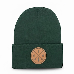 May include: A dark forest green knit beanie with a folded cuff. Centered on the cuff is a circular tan leather patch featuring a graphic of two crossed arrows, a small pine tree, and the letters N, P, and W positioned between the arrows.