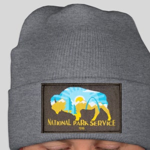 May include: A gray beanie featuring a patch with the National Park Service logo. The patch displays a yellow bison silhouette with a blue and yellow landscape, including mountains, trees, and a sunburst design. The text "NATIONAL PARK SERVICE 1916" is below the bison.