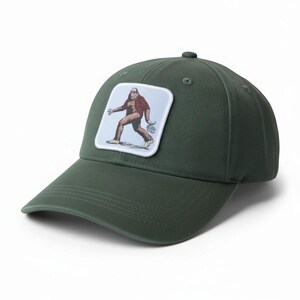 May include: A dark olive green baseball cap featuring a square patch on the front. The patch displays a light blue background with a graphic illustration of a brown, hairy creature wearing a red plaid shirt and a red cap, walking forward.