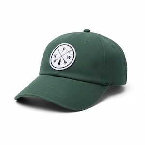 May include: A forest green baseball cap featuring a circular white patch on the front. The patch displays two crossed arrows and a small pine tree graphic, with the letters N, P, and W positioned between the arrows in a black circular border.