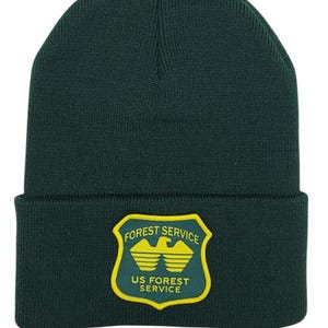 Pode incluir: Um gorro de malha verde escuro com um remendo amarelo e verde do Serviço Florestal dos Estados Unidos. O remendo apresenta uma águia com as asas abertas e as palavras "Forest Service" e "US Forest Service".