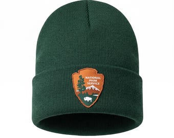 May include: A dark green knit beanie featuring a folded cuff. Centered on the cuff is an orange arrowhead-shaped patch with the text "NATIONAL PARK SERVICE" above a graphic of a tree, a mountain range, and a white bison silhouette.