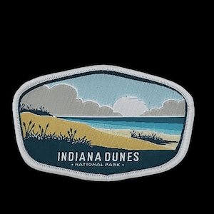May include: A blue and white embroidered patch featuring a landscape scene of a beach with sand dunes and a blue sky with clouds. The text "INDIANA DUNES NATIONAL PARK" is printed in white on a blue background.