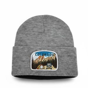 May include: A heather gray knit beanie featuring a rectangular patch on the front. The patch displays a colorful outdoor scene with mountains, a forest, a tent, and a vehicle, with the text COLORADO printed in bold blue letters at the top.