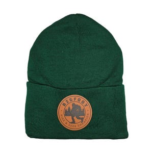Op de afbeelding: Een groene gebreide beanie met een bruine leren patch waarop in een cirkel "Bigfoot Hide & Seek Champion" staat.