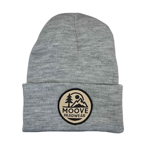 May include: A light gray knit beanie featuring a folded cuff and a circular patch on the front. The patch has a tan background with a black border and a black graphic depicting a pine tree, mountains, and a sun, with the text MOOVE HEADWEAR.