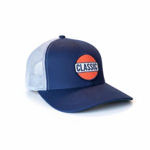 Könnte beinhalten: Marineblaue Trucker-Cap mit weißem Mesh-Rücken und gebogenem Schirm. Die Kappe hat einen runden Aufnäher mit dem Wort "CLASSIC" in weißen Buchstaben auf orangefarbenem Hintergrund.