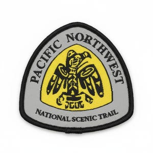 May include: A triangular embroidered patch with a grey border and background. It features a central yellow shield shape containing a black stylized bird totem graphic. The text PACIFIC NORTHWEST is arched above, and NATIONAL SCENIC TRAIL is below.