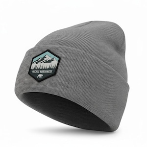 May include: A heather grey knit beanie with a folded cuff. Attached to the front is a black hexagonal patch featuring a light blue sky, white mountain peaks, a dark forest silhouette, a small white bear icon, and the text PACIFIC NORTHWEST.