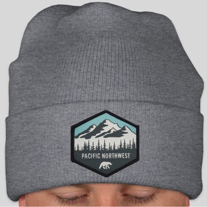 May include: Grey knitted beanie with a hexagonal patch featuring a mountain range, trees, and a bear. The patch has the text "PACIFIC NORTHWEST" on it.