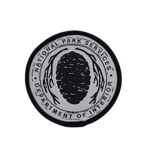 May include: A black and white embroidered patch with the text "National Park Service Department of Interior" in a circle around a stylized tree with a black and white pine cone.