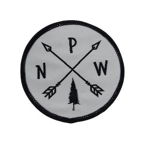 Pode incluir: Etiqueta bordada em preto e branco com um design de bússola. A bússola apresenta duas flechas apontando em direções opostas, um pinheiro e as letras "N", "P" e "W".