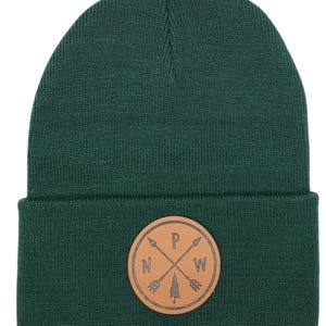 Könnte beinhalten: Eine dunkelgrüne Strickmütze mit einem braunen Lederpatch, der ein Kompassdesign mit Pfeilen zeigt, die nach Norden, Süden, Osten und Westen zeigen. Der Patch enthält auch eine kleine Grafik eines Kiefernbaums.