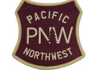 May include: A shield-shaped embroidered patch with a dark maroon background and a cream-colored border. The text PACIFIC is arched at the top, PNW is centered with a tree silhouette inside the N, and NORTHWEST is arched at the bottom in cream.