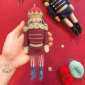 Könnte beinhalten: Zwei gehäkelte Nussknacker-Puppen, eine wird in der Hand gehalten. Die Puppen sind bordeauxrot und marineblau mit goldenen Akzenten. Sie haben rot-weiß gestreifte Beine und blaue Stiefel. Eine Schere und Wollknäuel sind ebenfalls zu sehen.