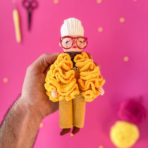 Może przedstawiać: Lalka szydełkowa w białym kapeluszu, czerwonych okularach, żółtej falbaniastych kurtce i brązowych spodniach. Lalka jest trzymana przez rękę na różowym tle.