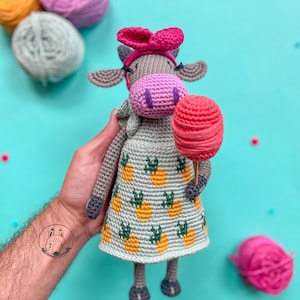 May include: A crocheted cow doll wearing a pink dress with a pineapple pattern and a pink bow. The doll is holding a pink yarn ball on a stick. There are balls of yarn in the background.