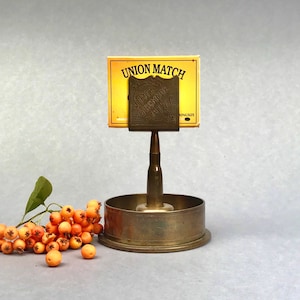 May include: A vintage match holder with a brass finish, holding a "UNION MATCH" box. The holder has a unique design, with a bullet-shaped stem and a circular base. A sprig of orange berries adds a natural touch.