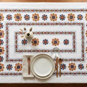 May include: White rectangular tablecloth with a detailed floral pattern in red, blue, and yellow. The tablecloth is set with a plate, cutlery, and a napkin. A small vase with dried flowers is also on the table.