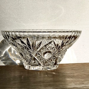 May include: A clear, cut-glass bowl with a wide, flared shape. The bowl features intricate geometric patterns and a starburst design at the base. The rim has a scalloped edge. The bowl sits on a wooden surface.